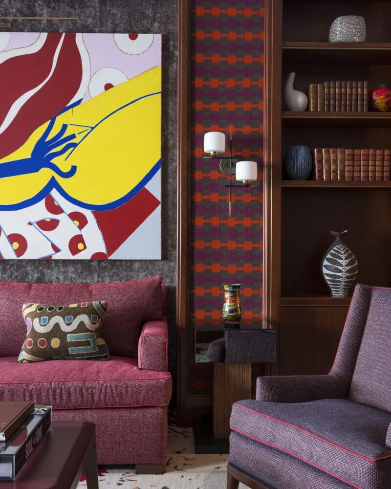 living room with big abstract painting above a purple couch with a brown coffee table, light purple love seat, wooden bookshelf, and brown and purple and orange striped wallpaper